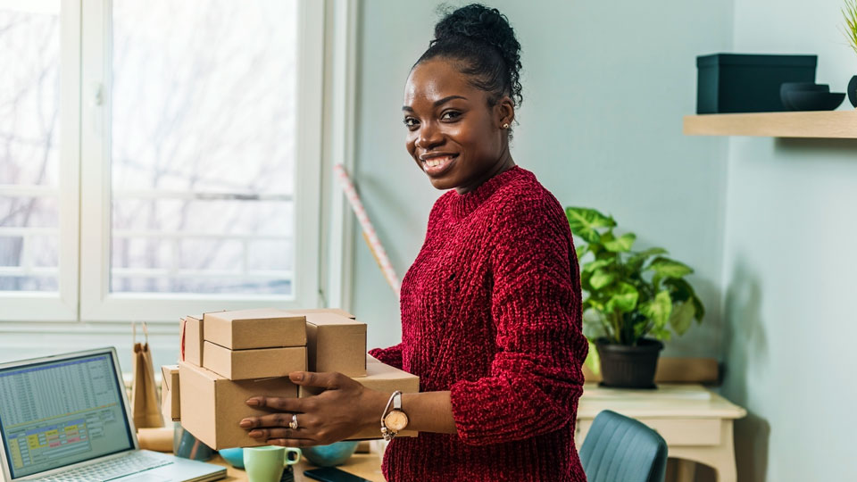 Female small business owner holding packages in her workspace smiling.