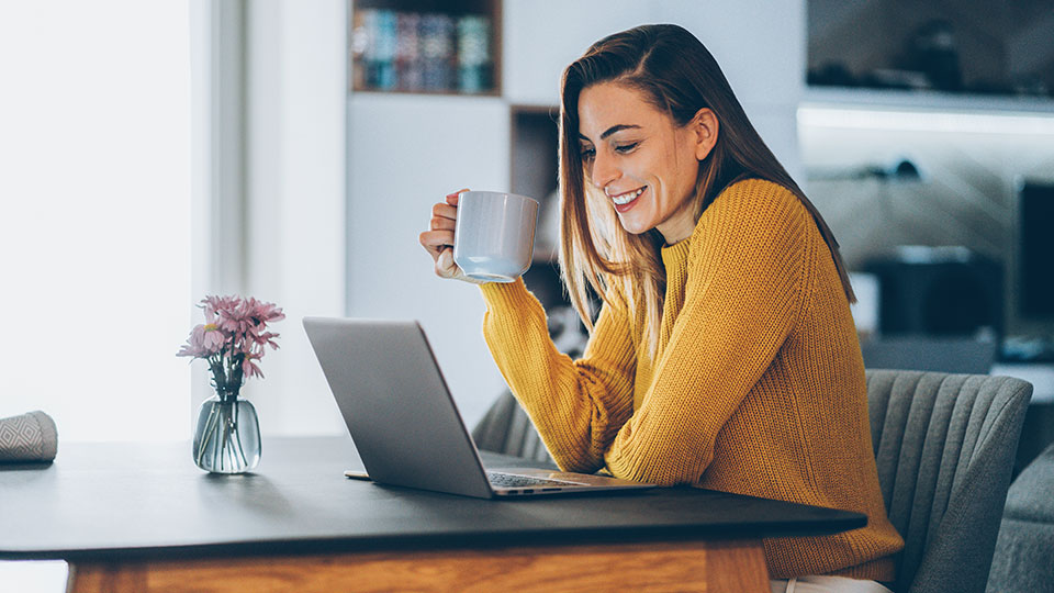 Woman sitting looking at laptop while holding a coffee mug in her hand.